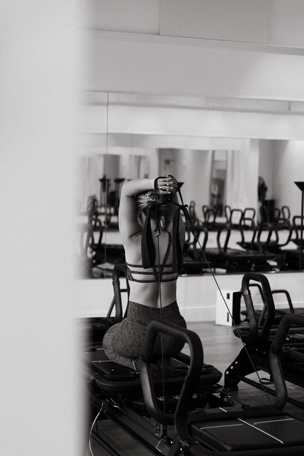 Person stretching arms behind head while seated on a Pilates reformer machine in a studio.