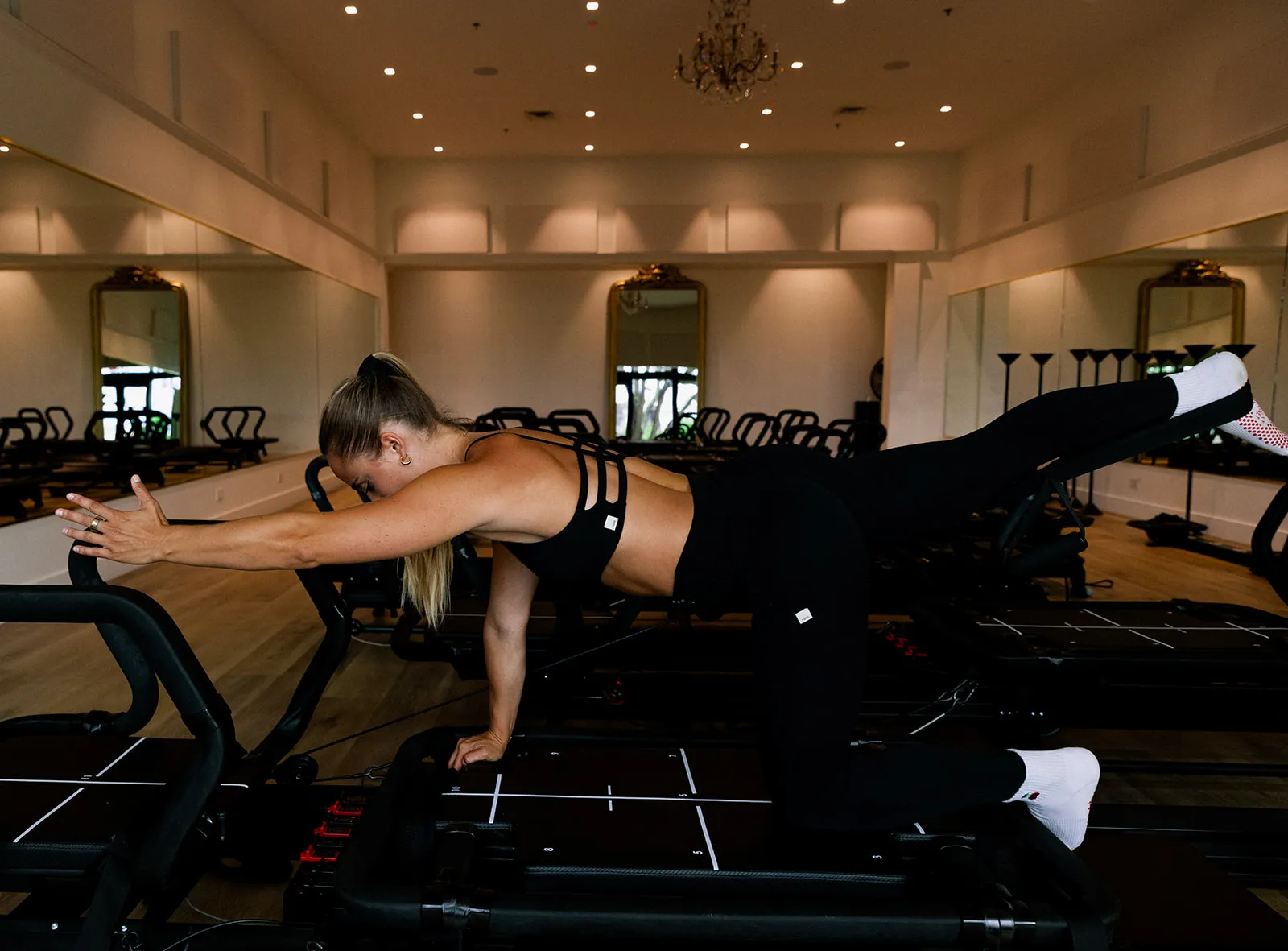 Woman in black workout clothes stretching arm and leg on Pilates reformer machine in studio.