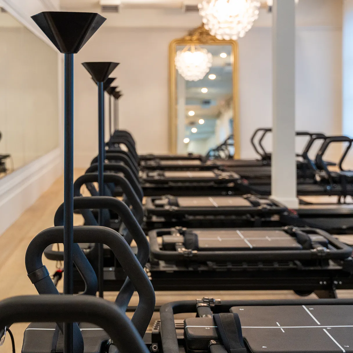 Row of black Pilates reformer machines lined up in a bright studio with mirrors and chandeliers.