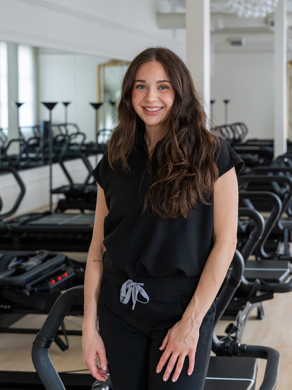 Smiling woman with long brown hair wearing black workout clothes standing in a Pilates studio with reformer machines.
