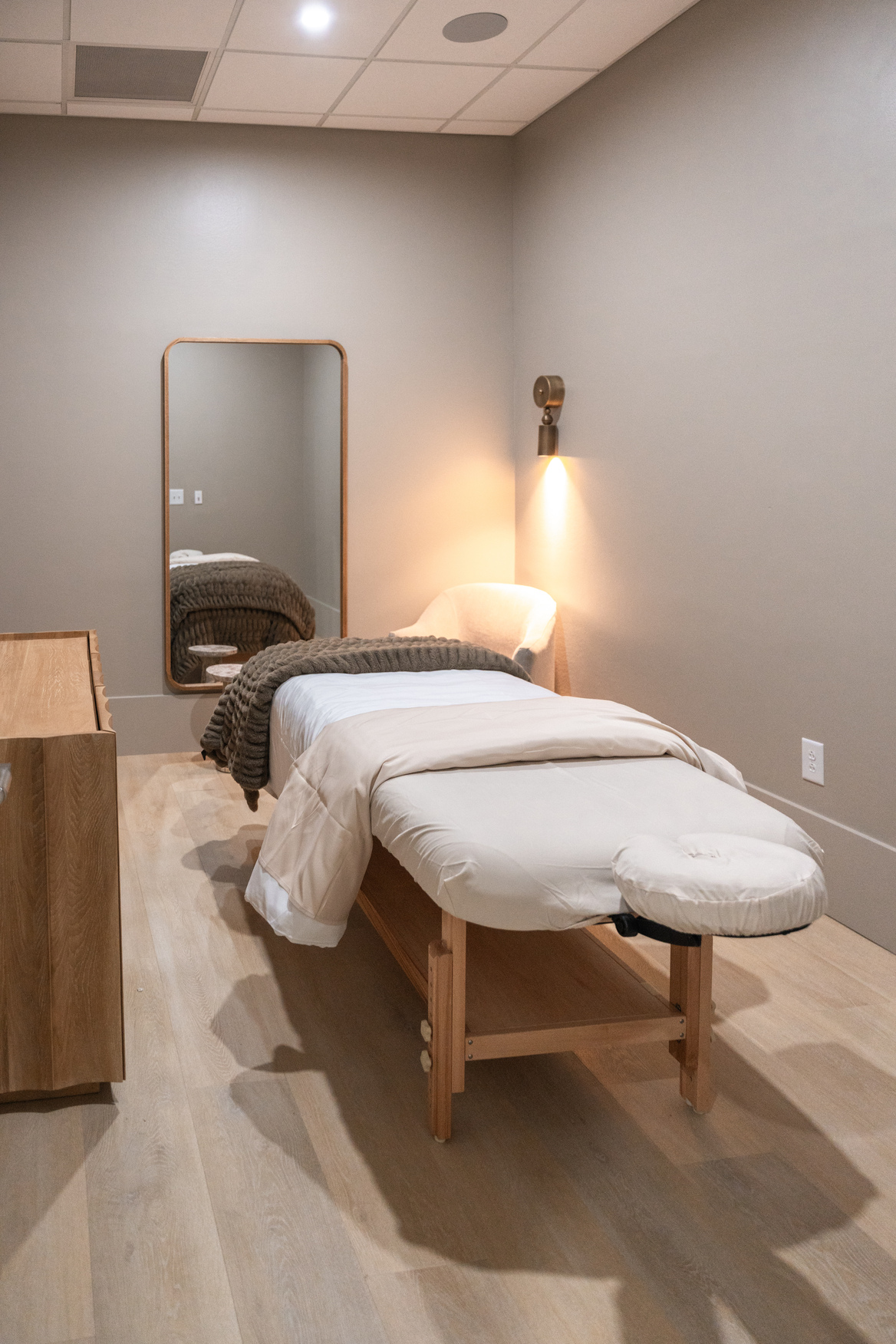 Calm massage therapy room with an empty massage table, a blanket, a chair, a wooden cabinet, and a wall mirror.