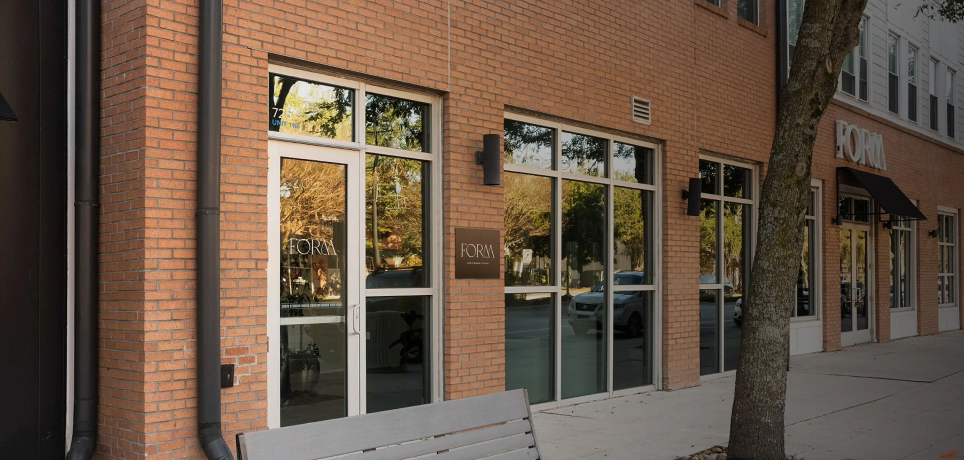 Brick building with large windows and two doors, a tree, and a bench on the sidewalk.