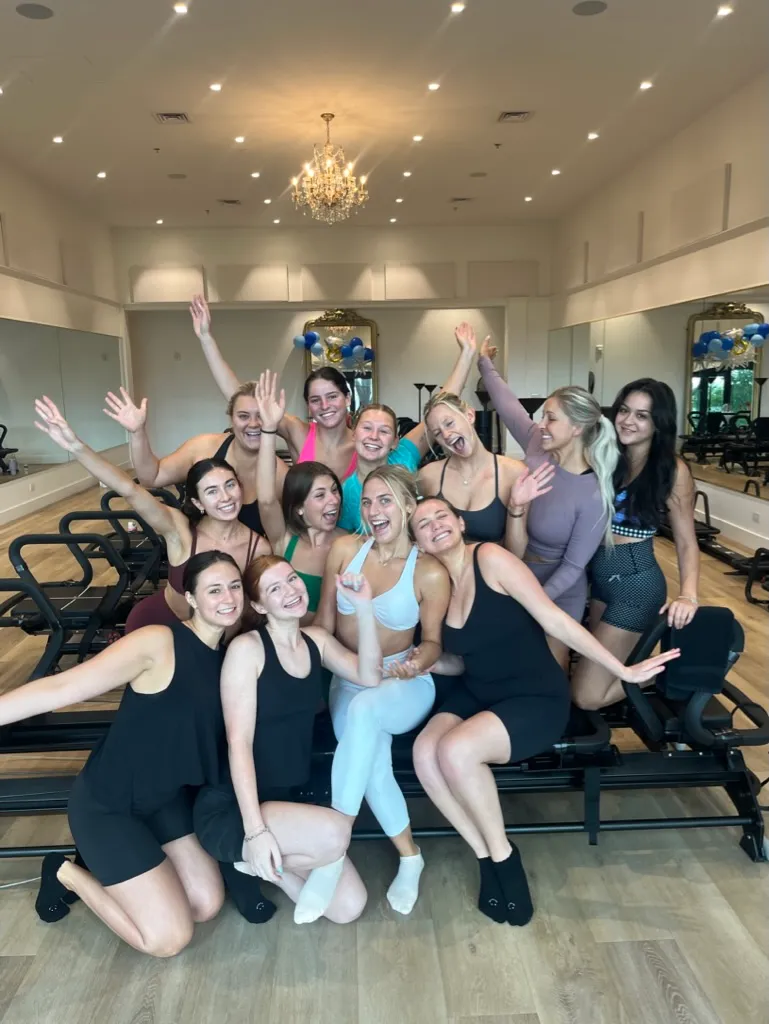 Group of women in athletic wear posing happily on Pilates reformer machines in bright studio.