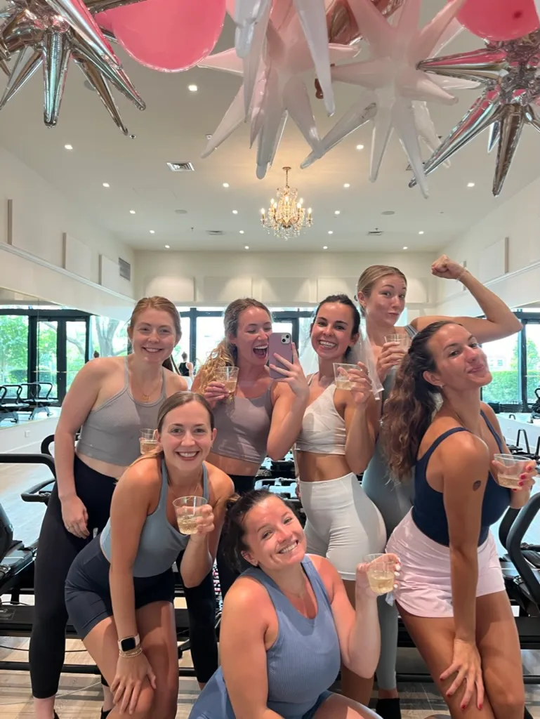 Seven women in workout clothes smiling and holding drinks in a gym with exercise bikes and chandelier.