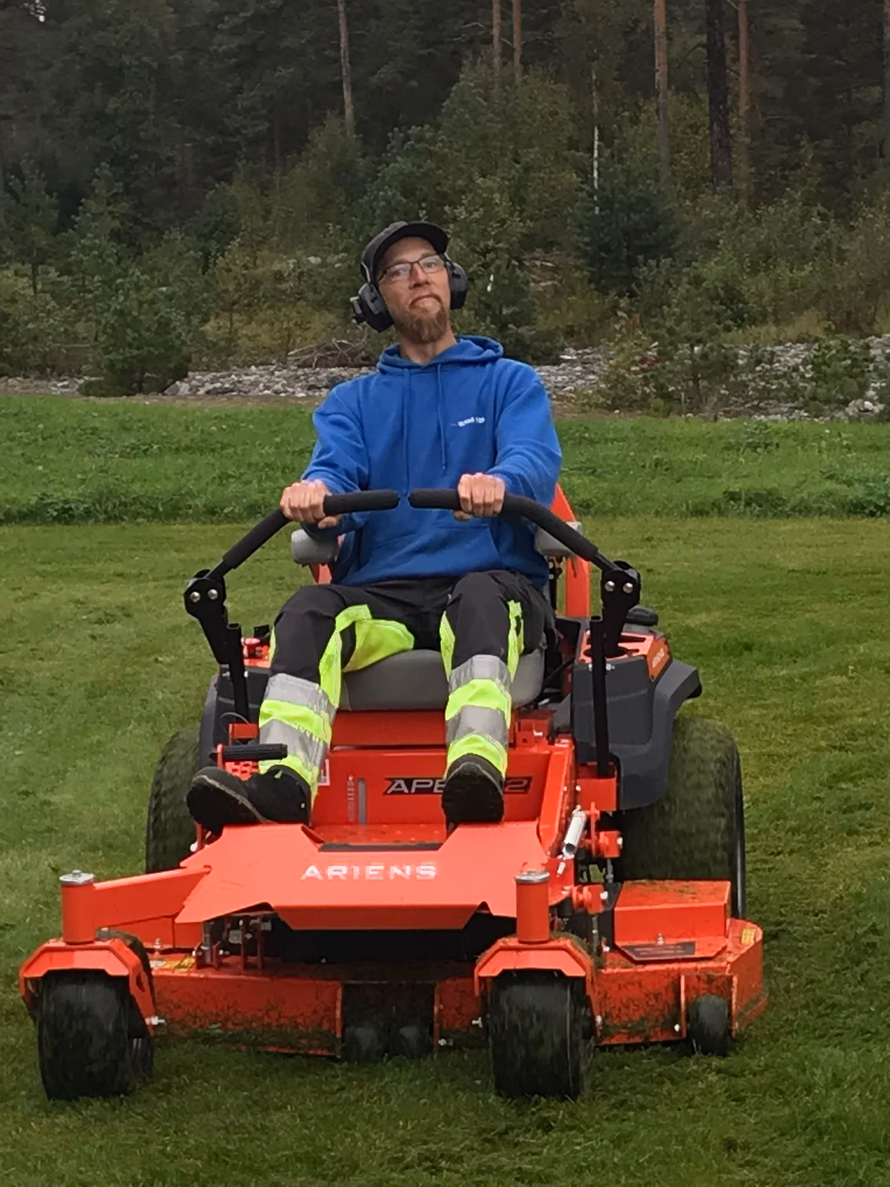 Mann i blå hettegenser og refleksbukser kjører oransje Ariens gressklipper på en grønn plen med skog i bakgrunnen.