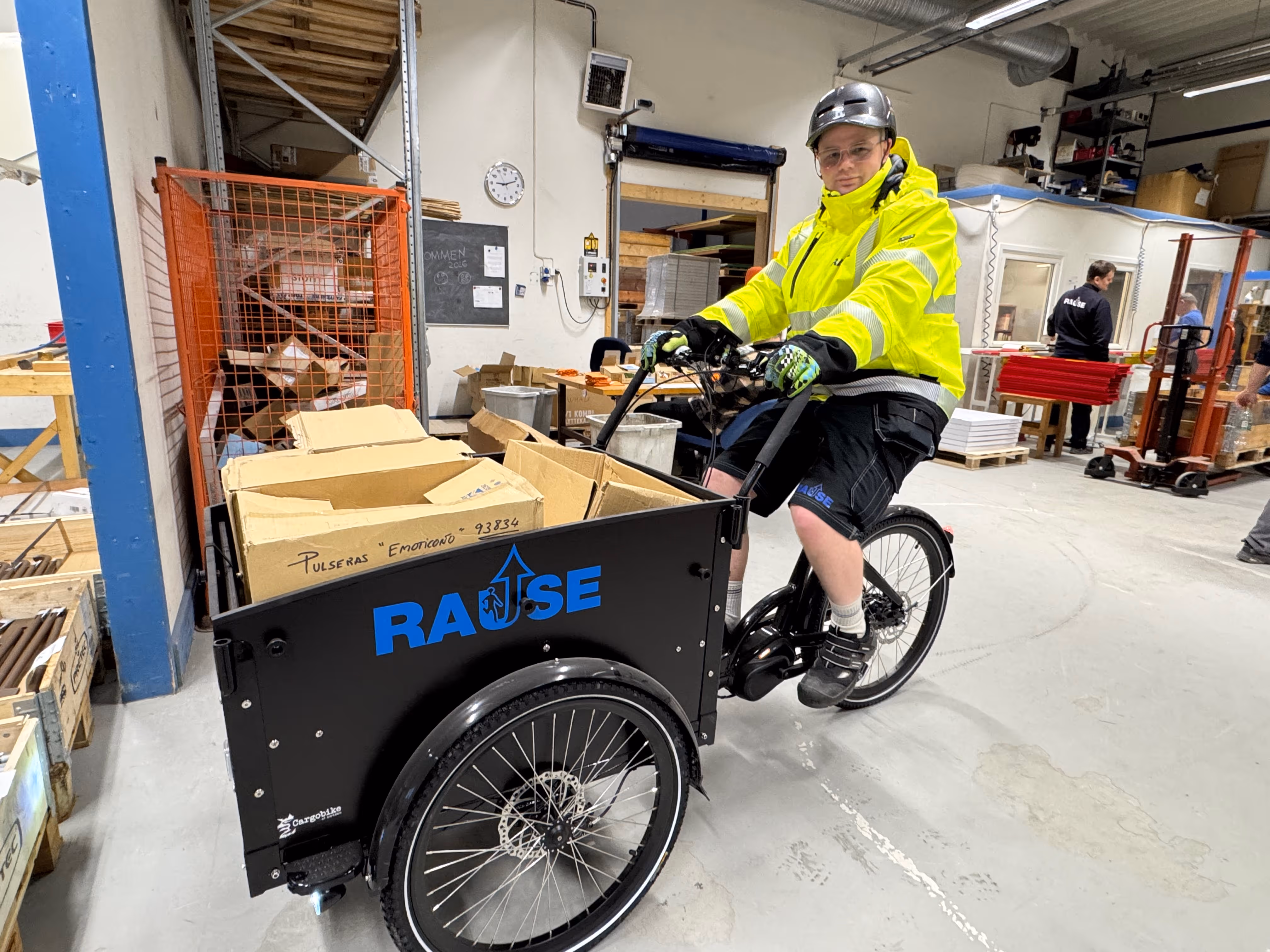 Person med gul refleksjakke og hjelm sitter på en trehjulssykkel med kasse lastet med pappesker i et lagerbygg.