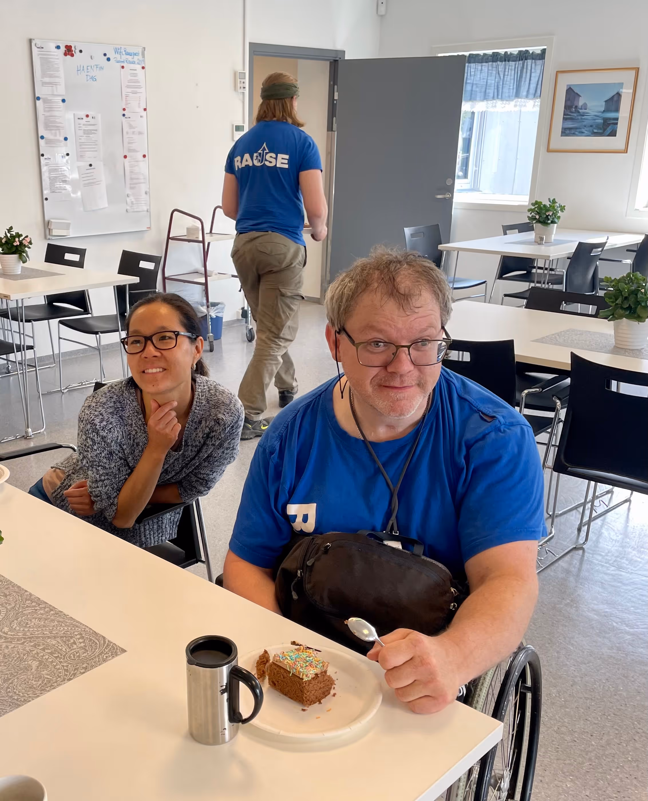 To personer i et lyst rom med spisebord, en mann i rullestol med blå skjorte og briller som holder en skje ved et stykke sjokoladekake med strøssel på en tallerken.
