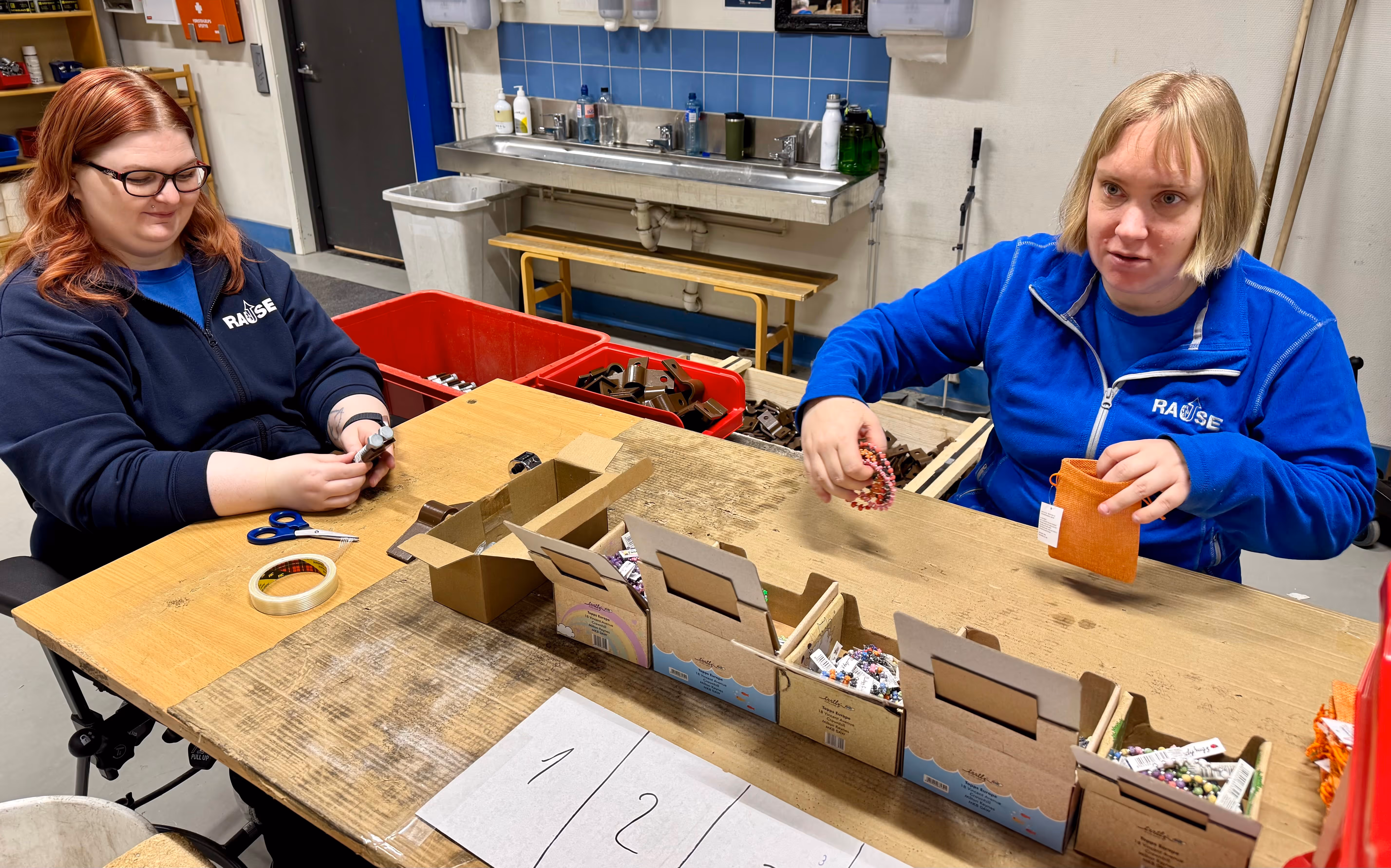 To kvinner sitter ved et bord og sorterer fargede perlearmbånd i esker merket med tall i et verksted.