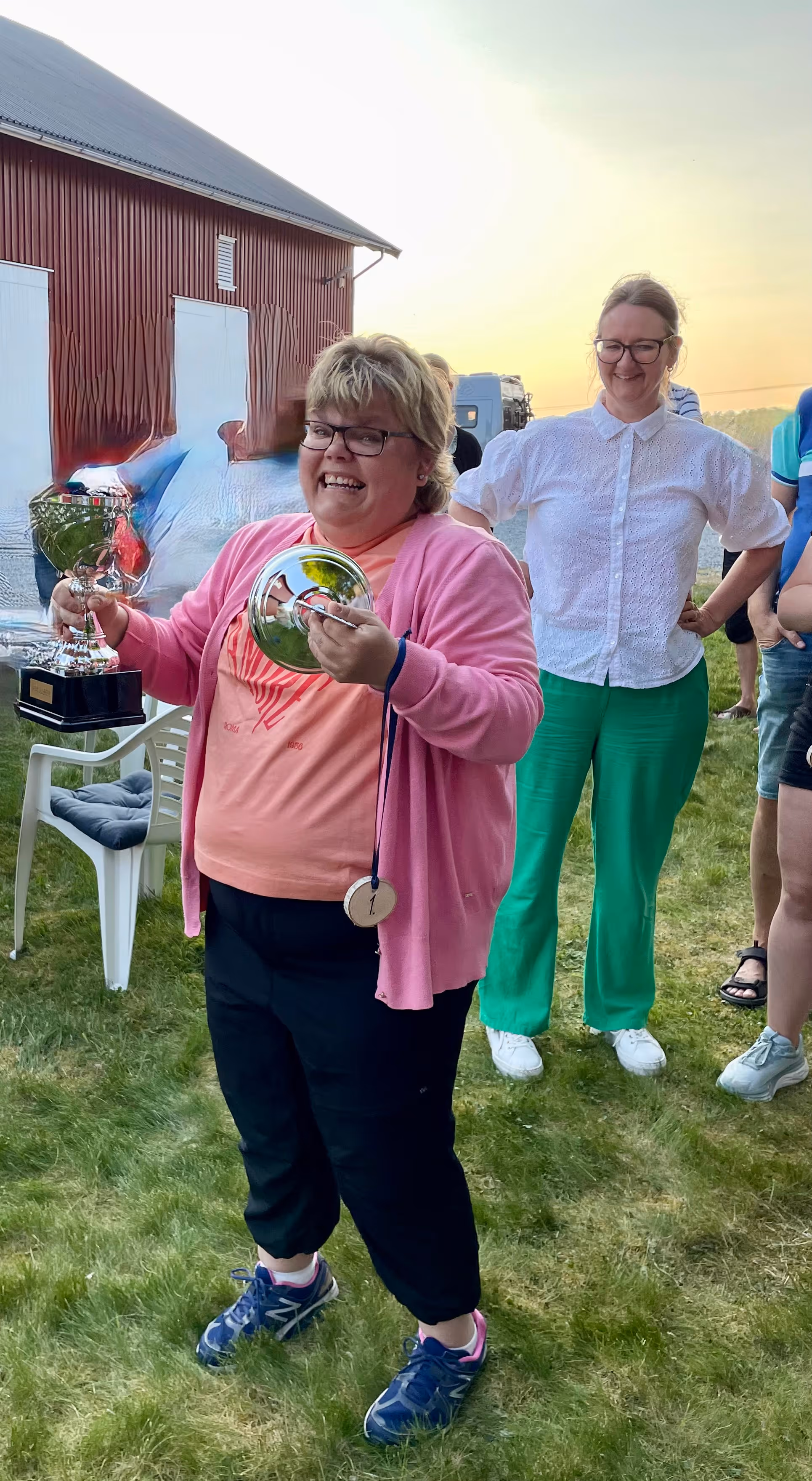 Kvinne iført rosa jakke og t-skjorte holder en pokal og et sølvfat med et vinnermerke på gress med flere mennesker og låve i bakgrunnen.