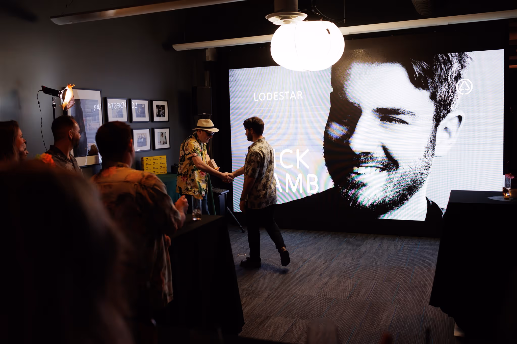 Two men shaking hands in a dimly lit room with a large screen showing a black and white portrait and text behind them.