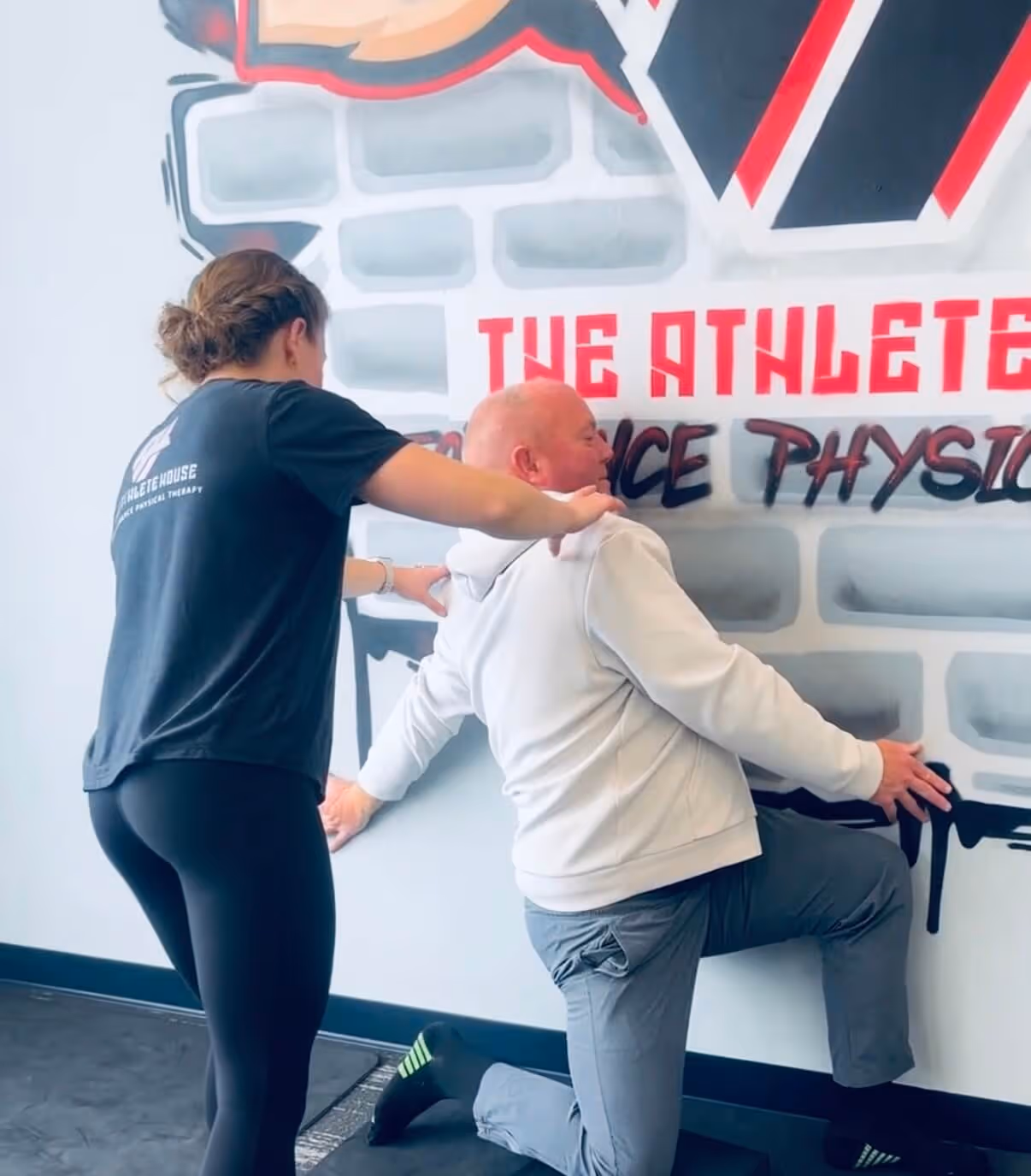 Physical therapist assisting a man in a white hoodie with a knee stretch against a graffiti wall inside a therapy clinic.