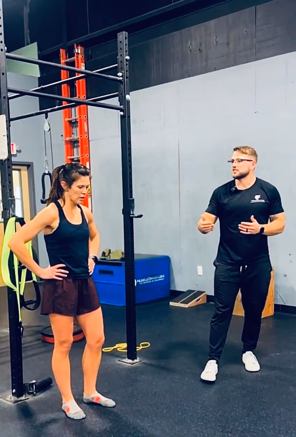 Woman in workout attire standing with hands on hips listening to a man in black athletic clothing speaking in a gym.
