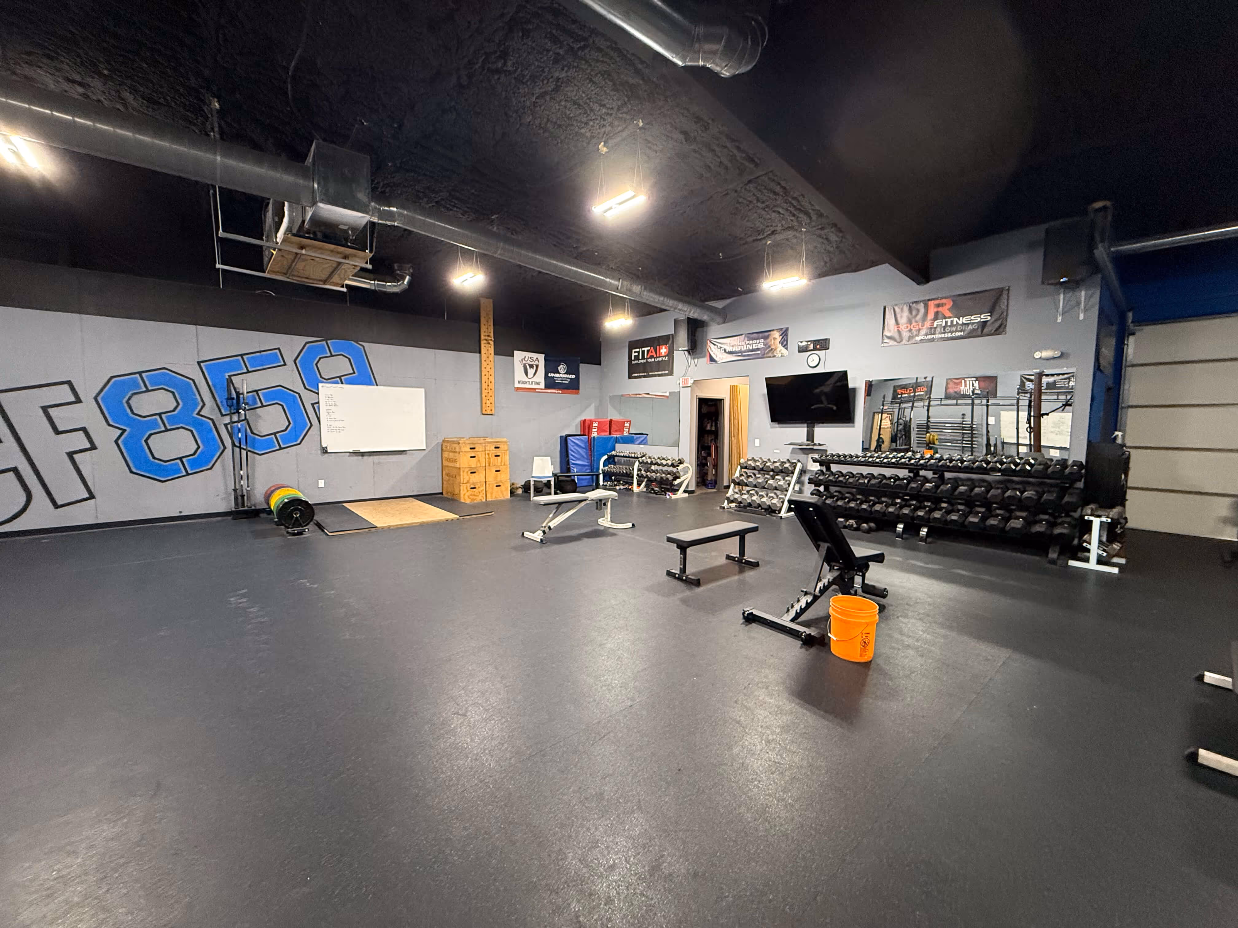 Indoor gym space with benches, dumbbell racks, weightlifting platform, wall-mounted TV, and large SF859 logo on the wall.