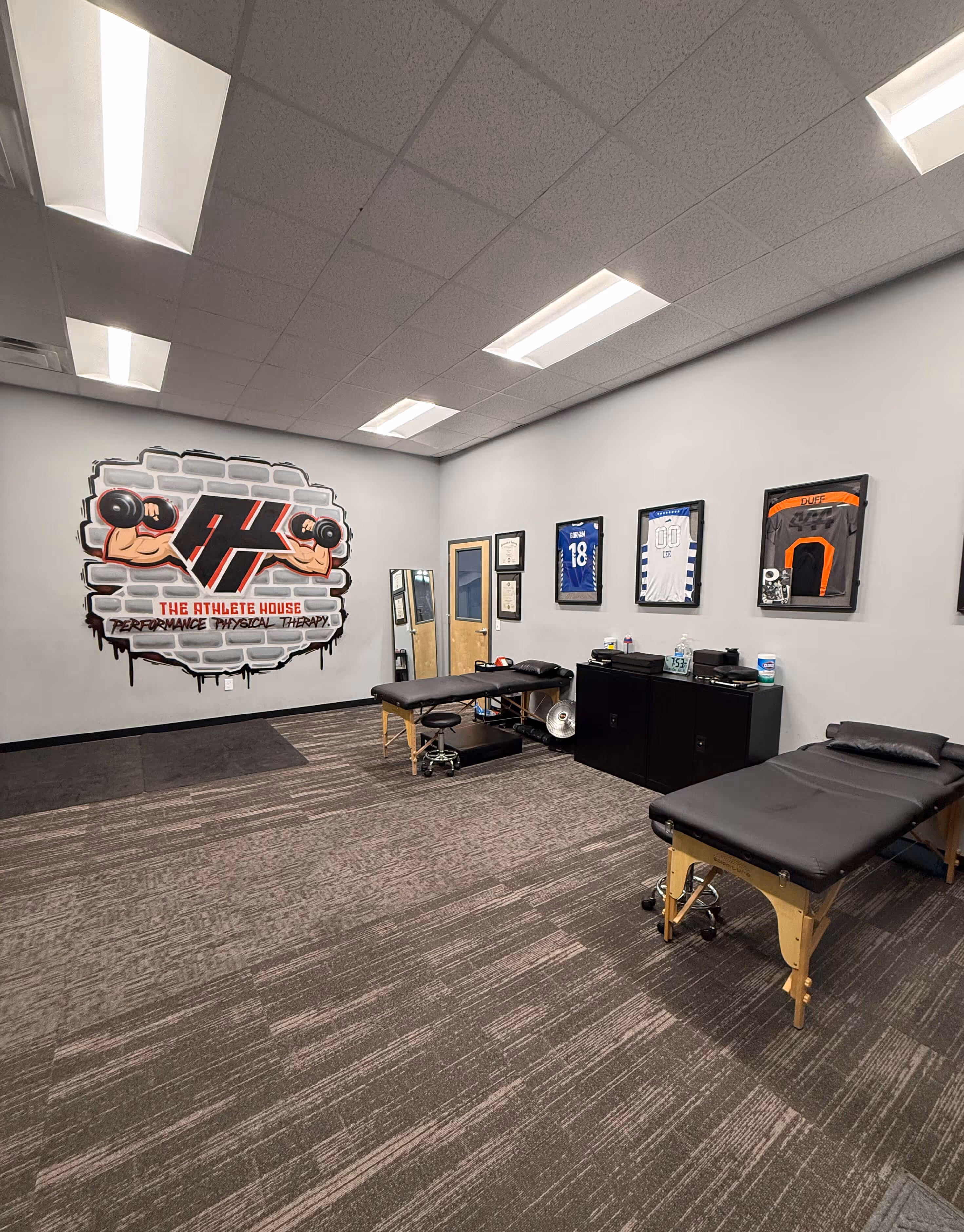 Interior of a physical therapy room with two black treatment tables, wall art with dumbbells logo, framed sports jerseys, and certificates.