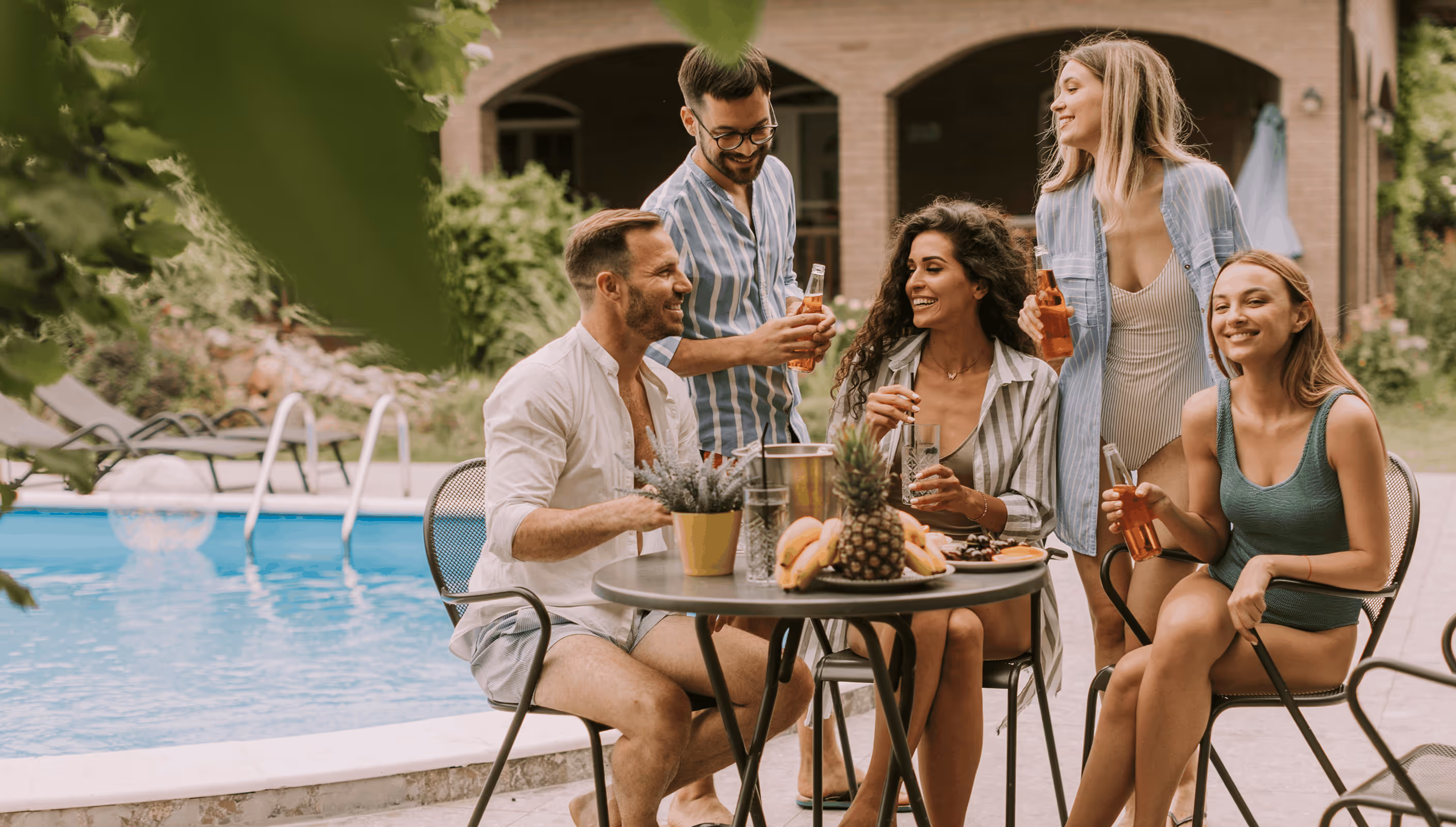 Group of five friends relaxing and drinking by a poolside table with fruit and plants.