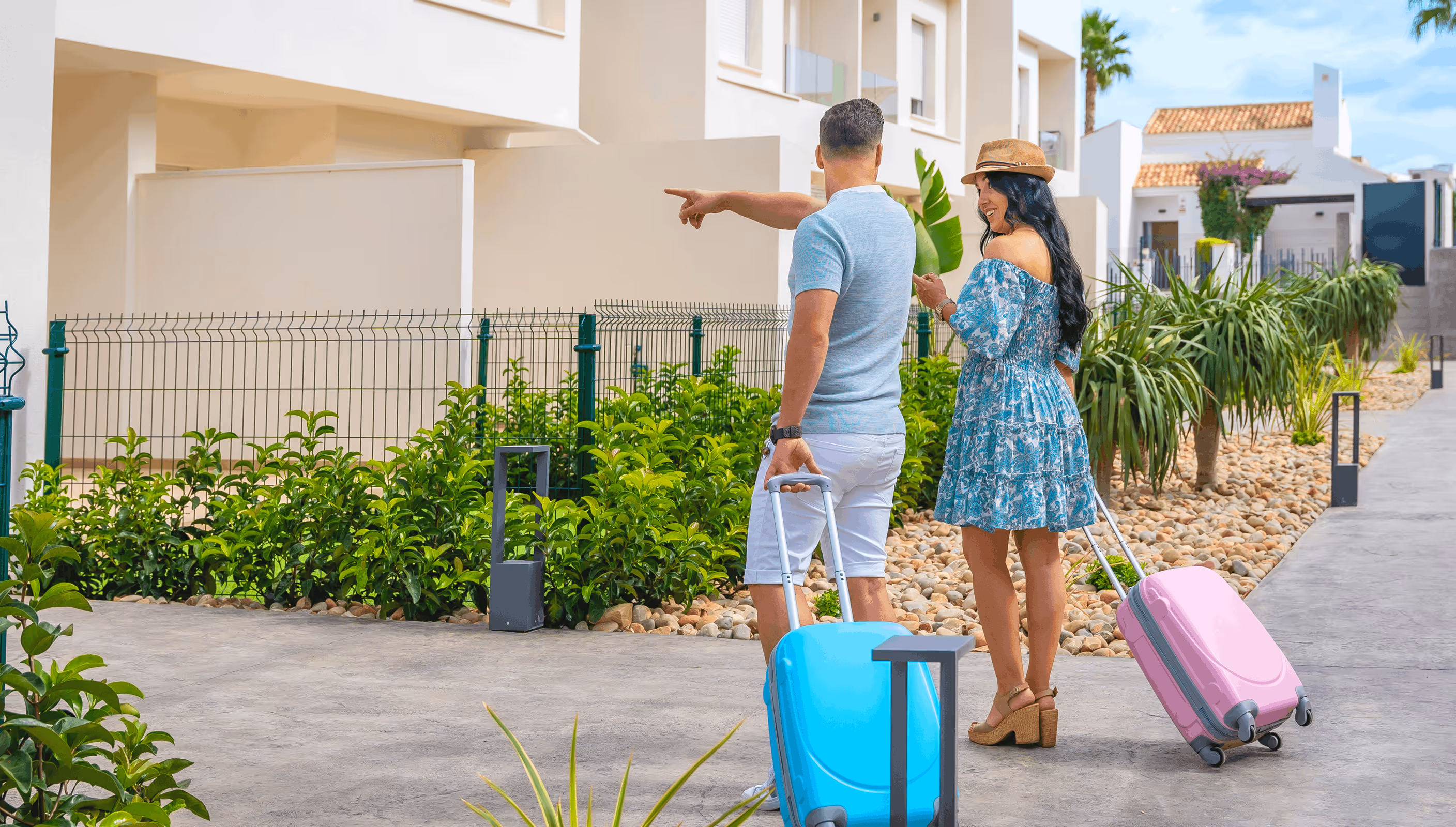 Couple pulling suitcases, walking outdoors near modern white buildings and greenery, with man pointing ahead.
