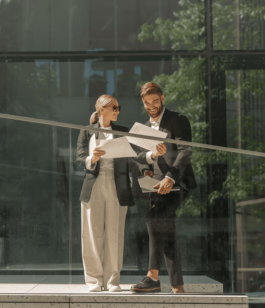 Two business professionals reviewing documents together while standing outdoors near a glass railing.