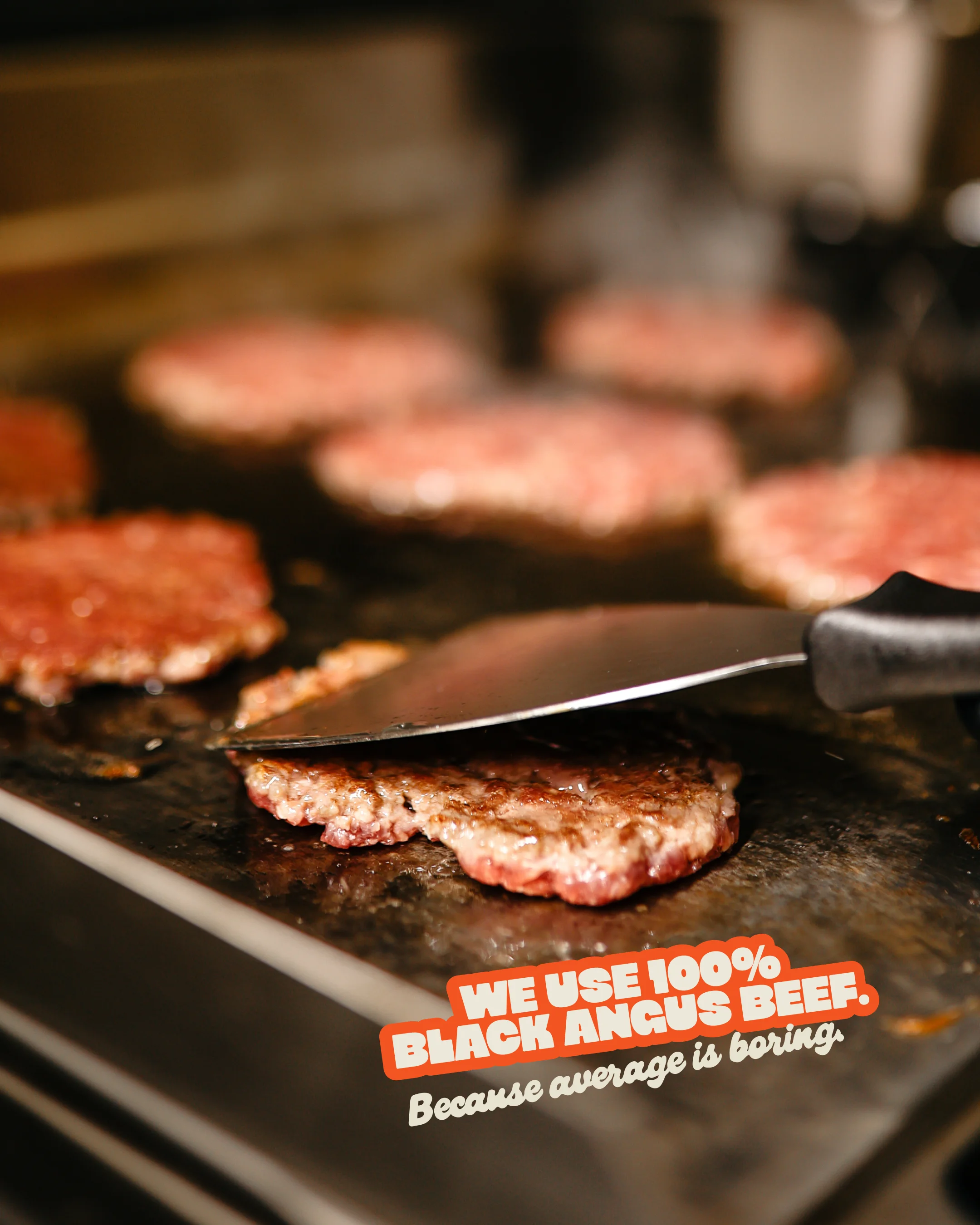 Close-up of multiple hamburger patties cooking on a flat grill with a spatula lifting one patty, text overlay states 'We use 100% Black Angus beef. Because average is boring.'