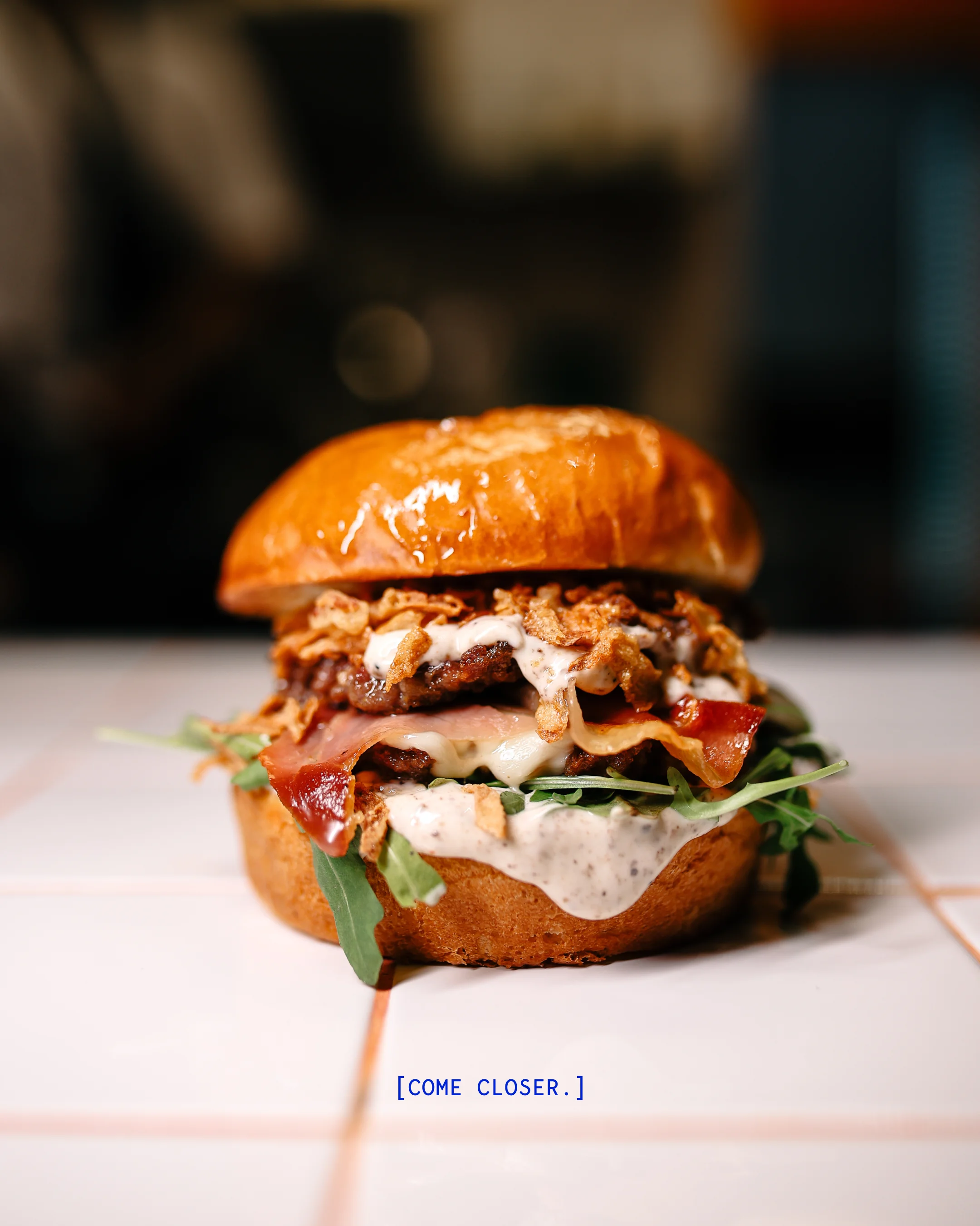 Close-up of a gourmet burger with crispy onions, bacon, cheese, arugula, and creamy sauce on a glossy bun.