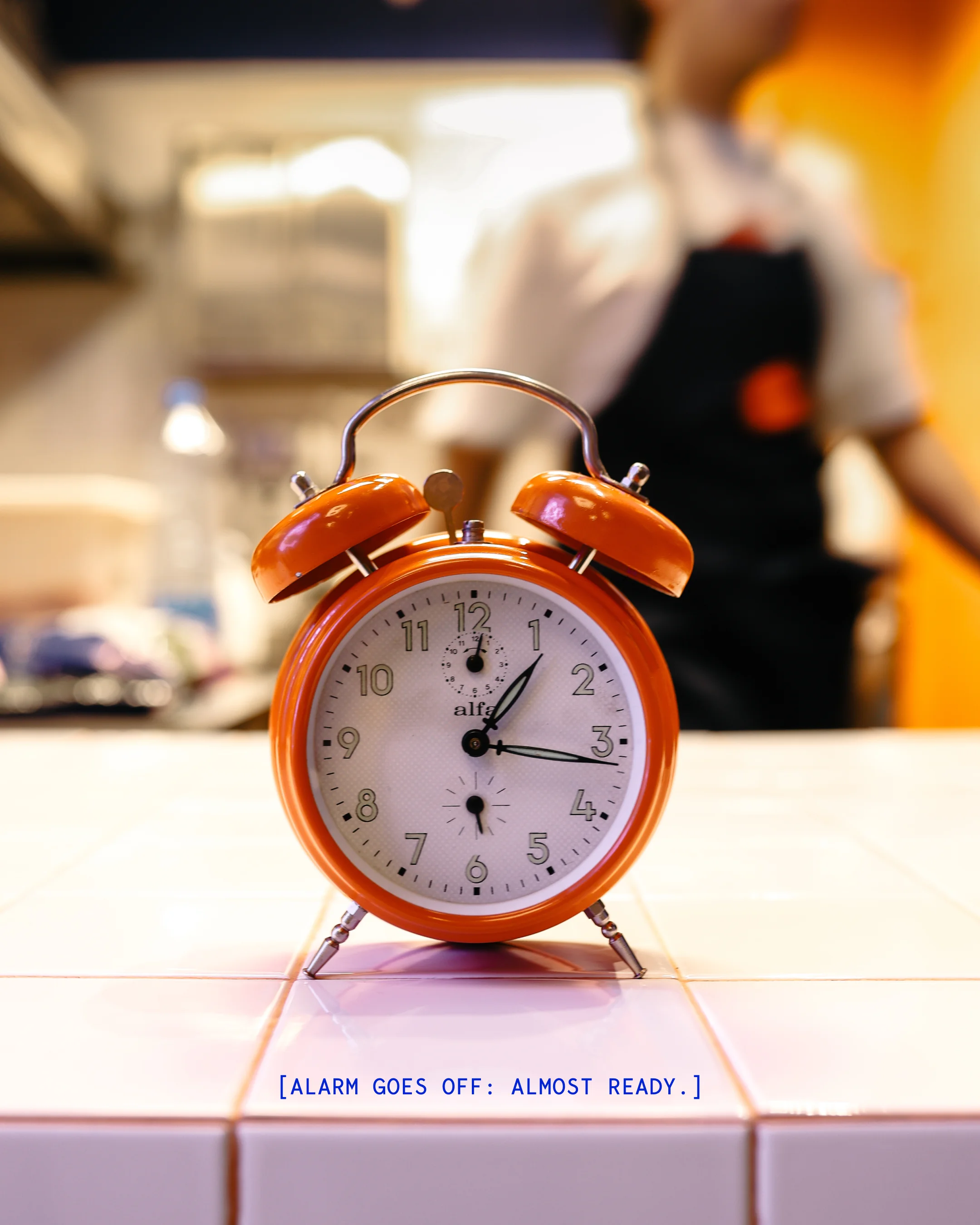 Orange twin-bell alarm clock on white tiled surface showing 1:15 with blurred background and text '[ALARM GOES OFF: ALMOST READY.]'