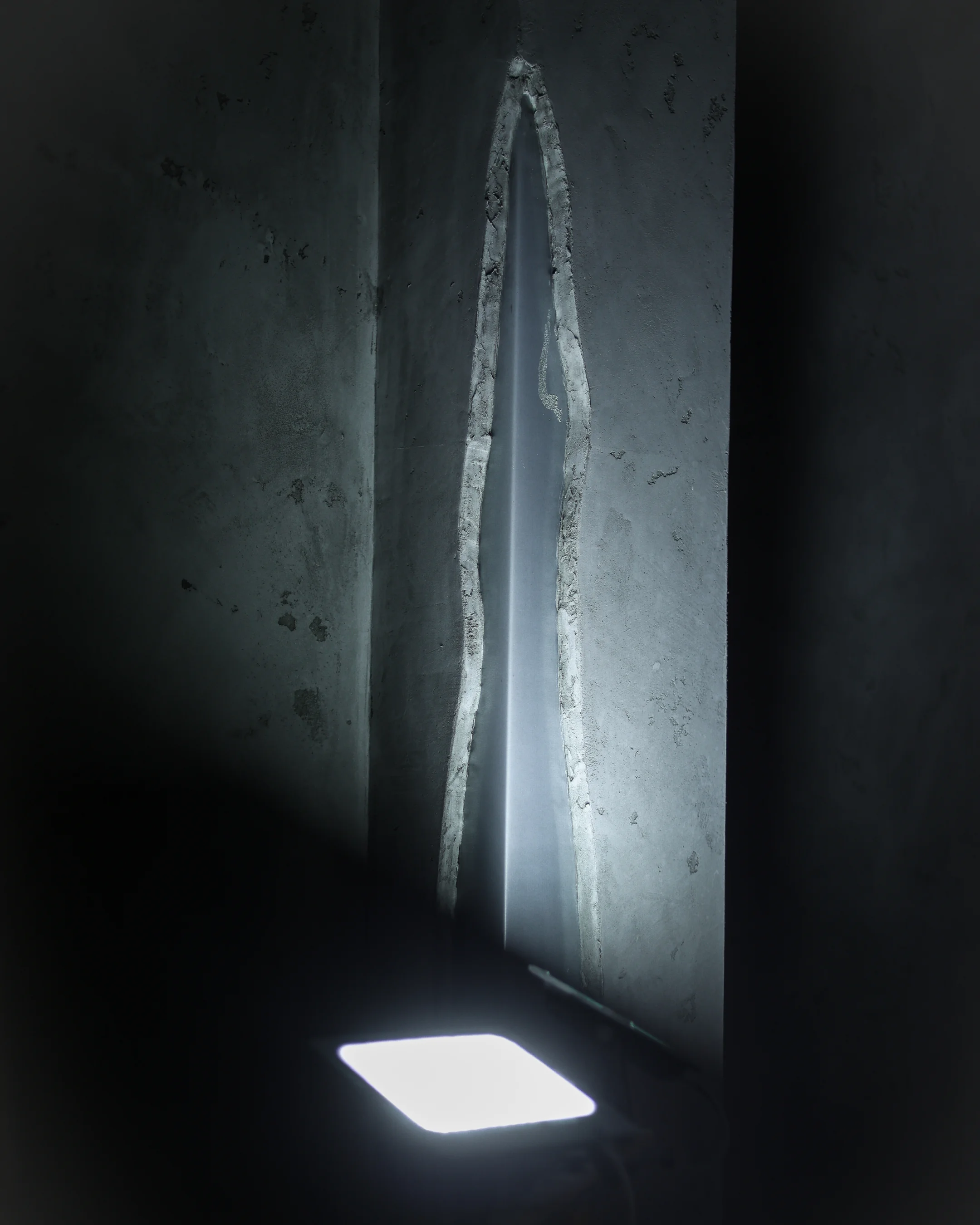 Crack in a concrete wall illuminated by a bright rectangular light source in a dark setting.