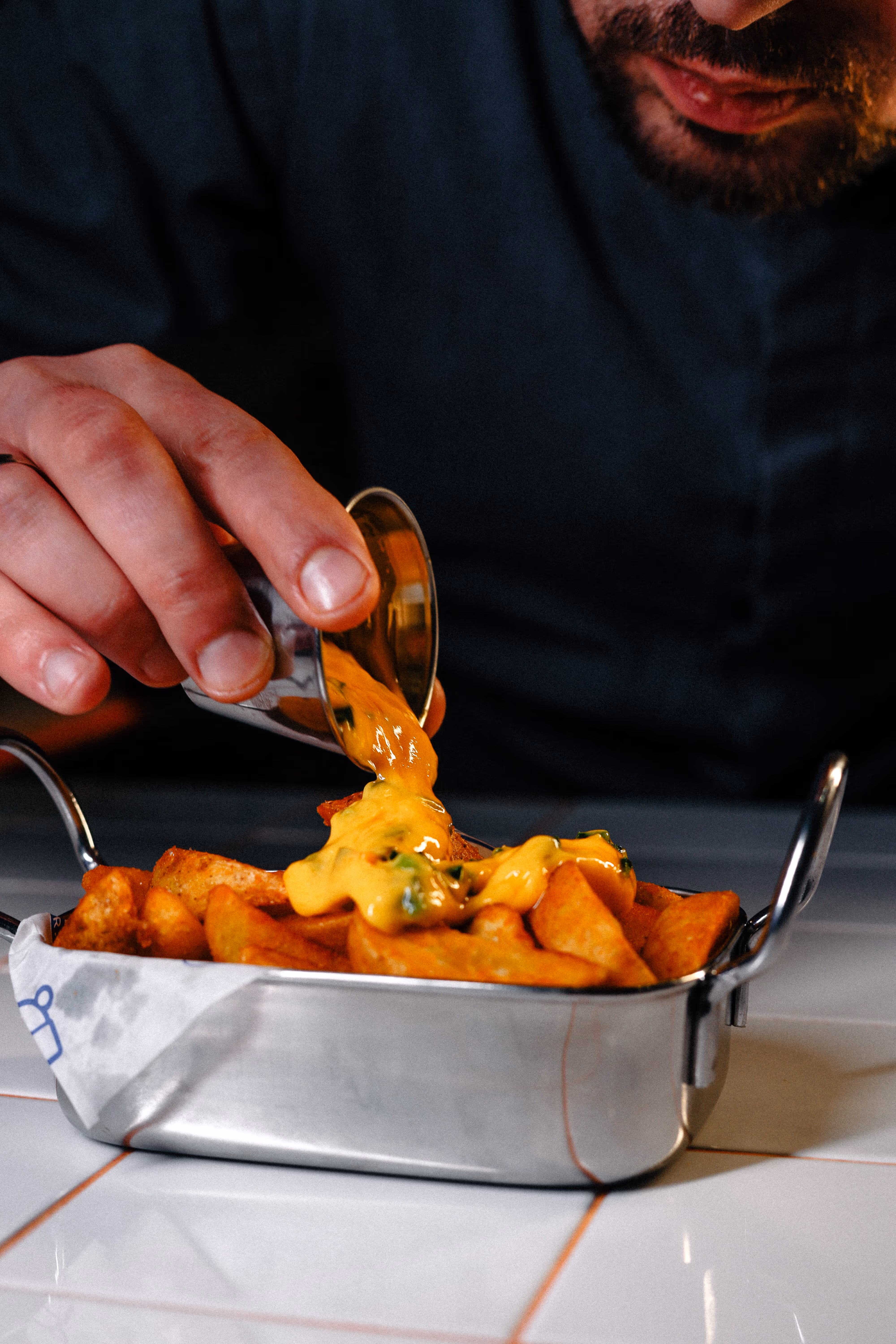 Person pouring melted cheese sauce over crispy potato wedges in a small metal serving tray.