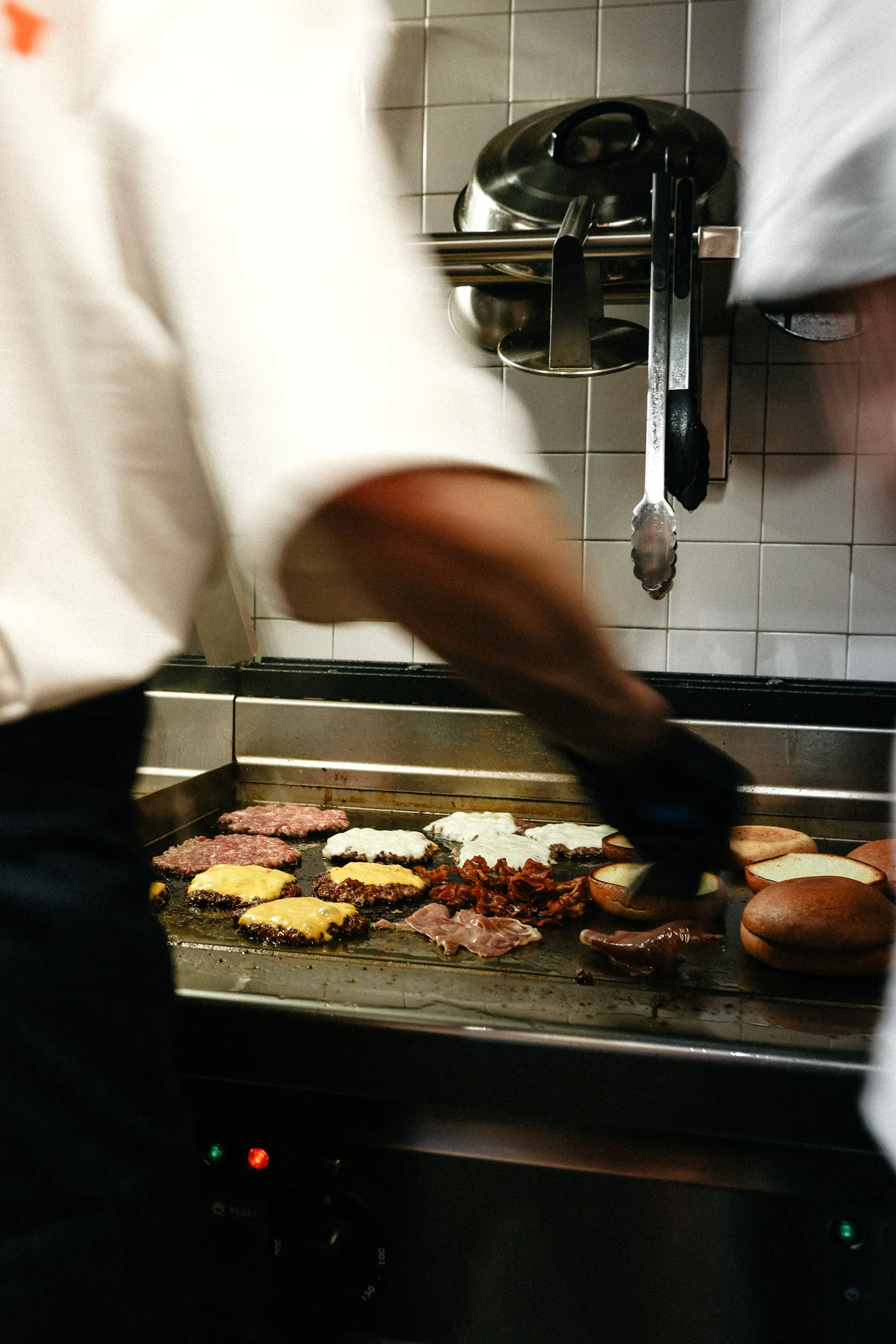 Chef cooking multiple cheeseburgers, bacon, and toasting buns on a large flat grill in a kitchen.