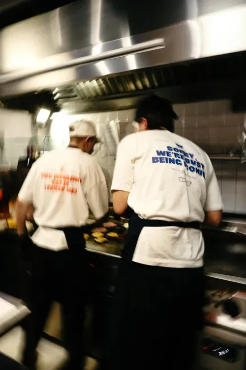 Two chefs cooking on a grill in a kitchen wearing white t-shirts with text on the back.