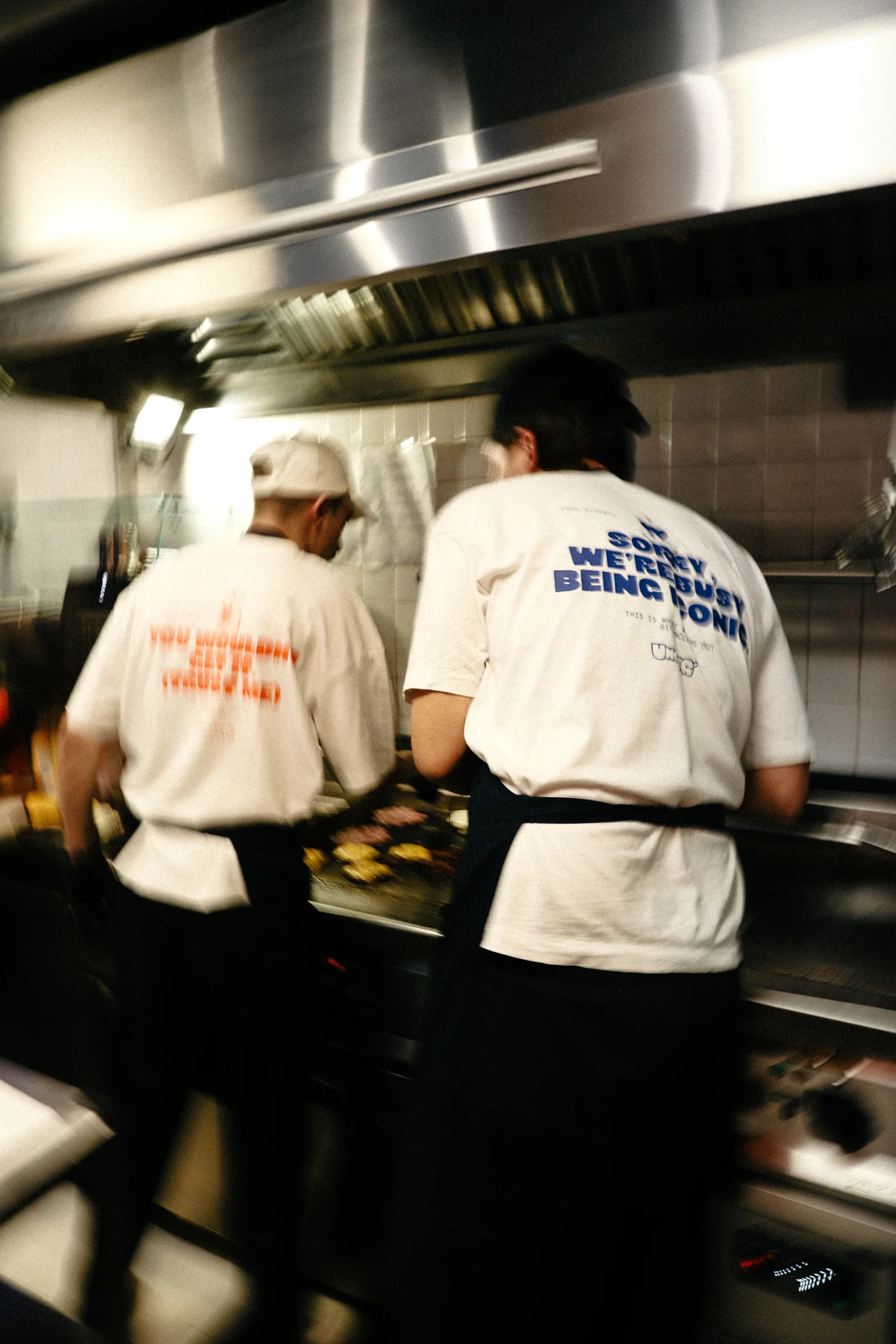 Two chefs cooking on a grill in a kitchen wearing white t-shirts with text on the back.