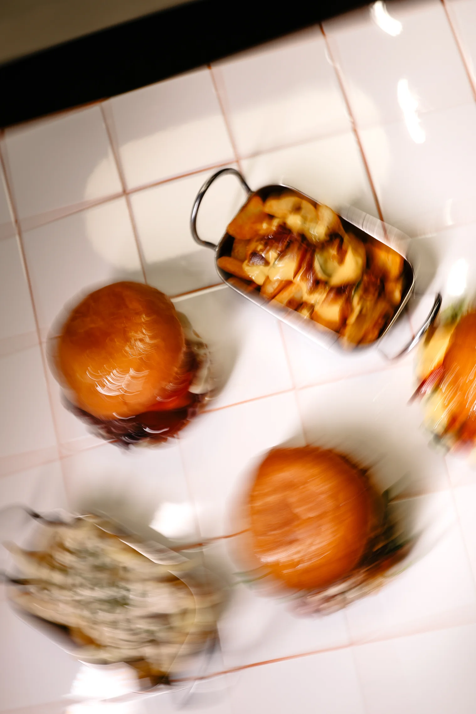 Blurred image of two burgers and two containers of loaded fries on a tiled surface.