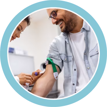 Healthcare professional drawing blood from a smiling man's arm with a green tourniquet in a clinical setting.