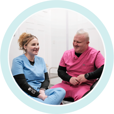 Two healthcare professionals smiling and sitting in a clinical setting, one wearing blue scrubs and the other in pink scrubs.