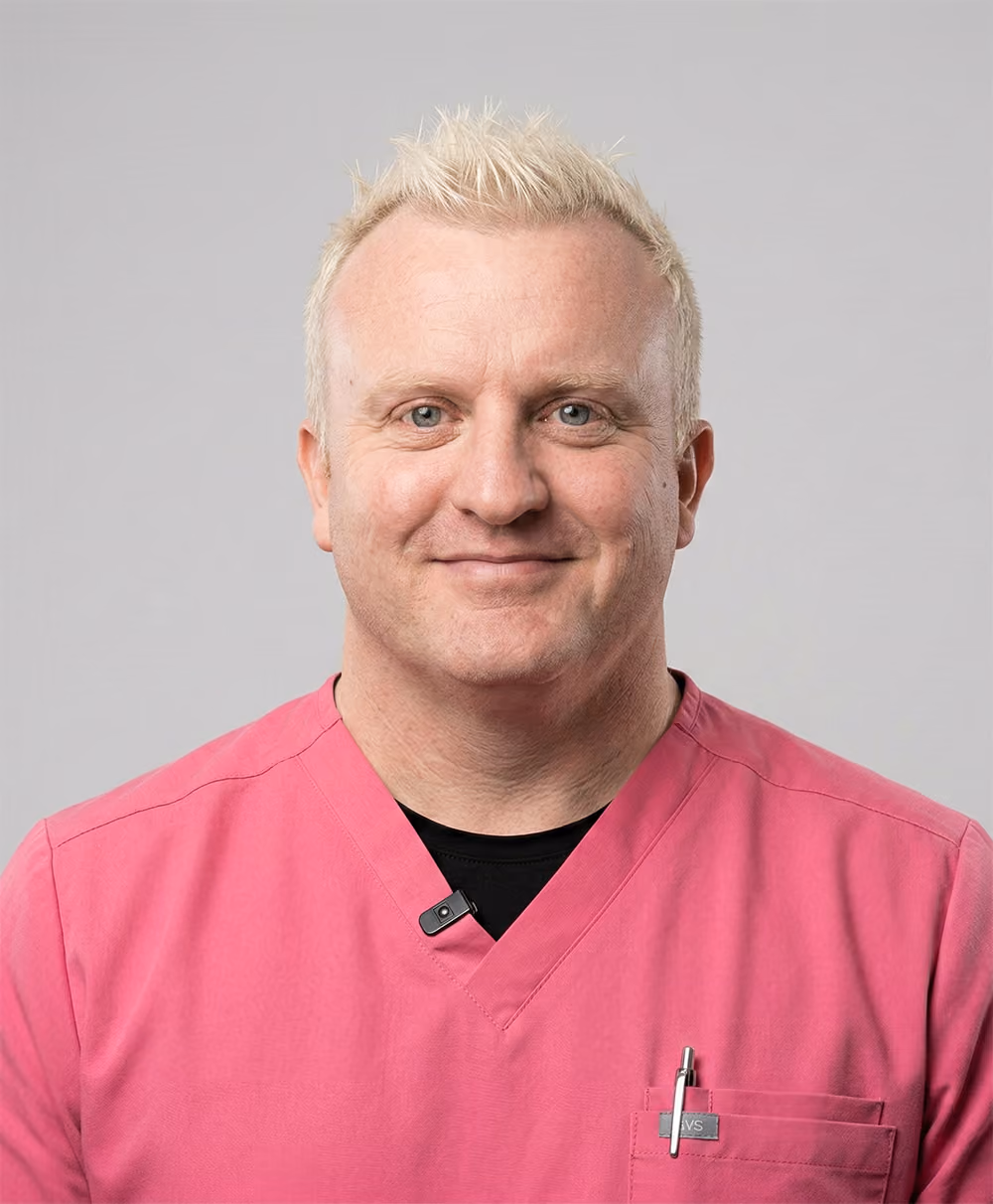Smiling man with short blond hair wearing a pink scrub top and a pen in his chest pocket against a plain light gray background.