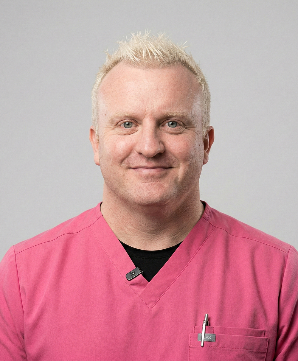 Smiling man with short blond hair wearing a pink scrub top and a pen in his chest pocket against a plain light gray background.