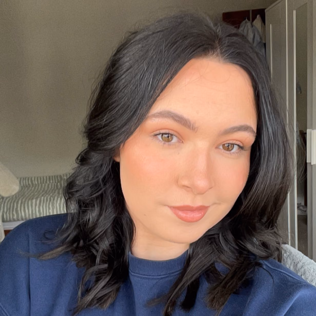 Young woman with dark wavy hair and light makeup wearing a navy blue top inside a room.
