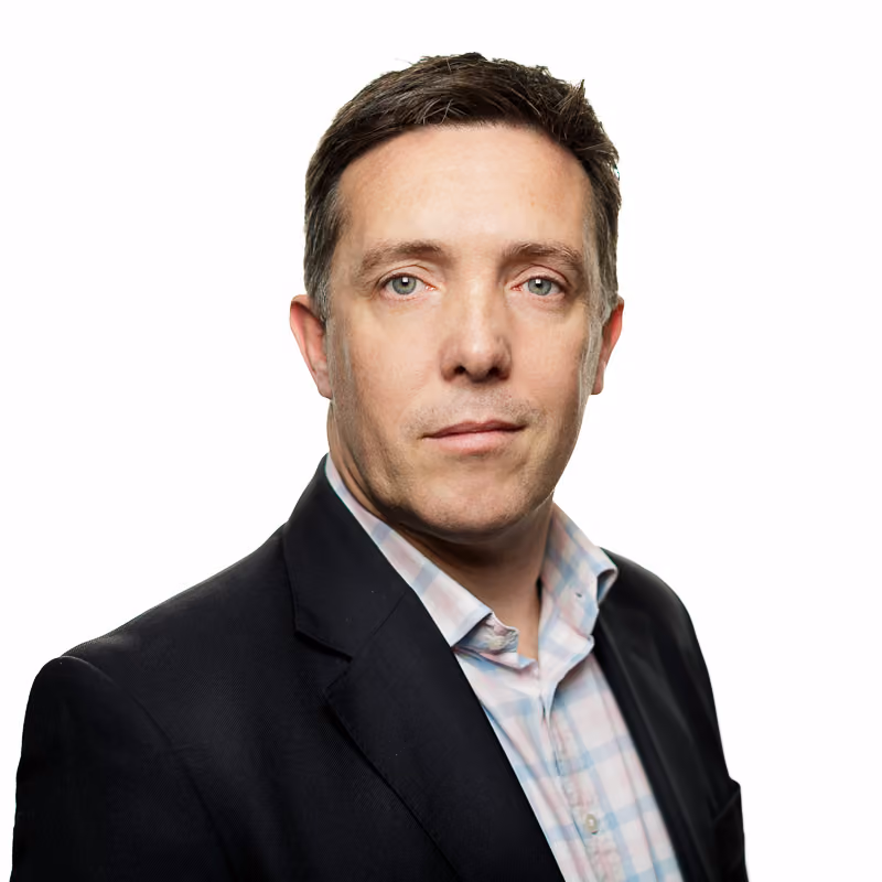 Man with short brown hair wearing black blazer and light plaid shirt on white background.