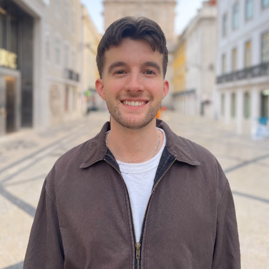 Young man with short dark hair and brown jacket smiling on a paved city street.