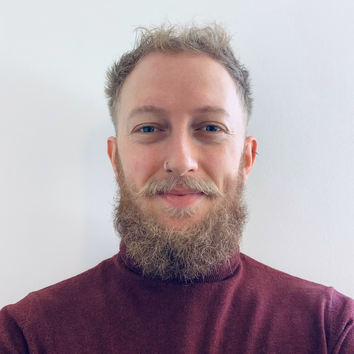 Man with curly beard and mustache wearing a maroon turtleneck smiling against a white background.