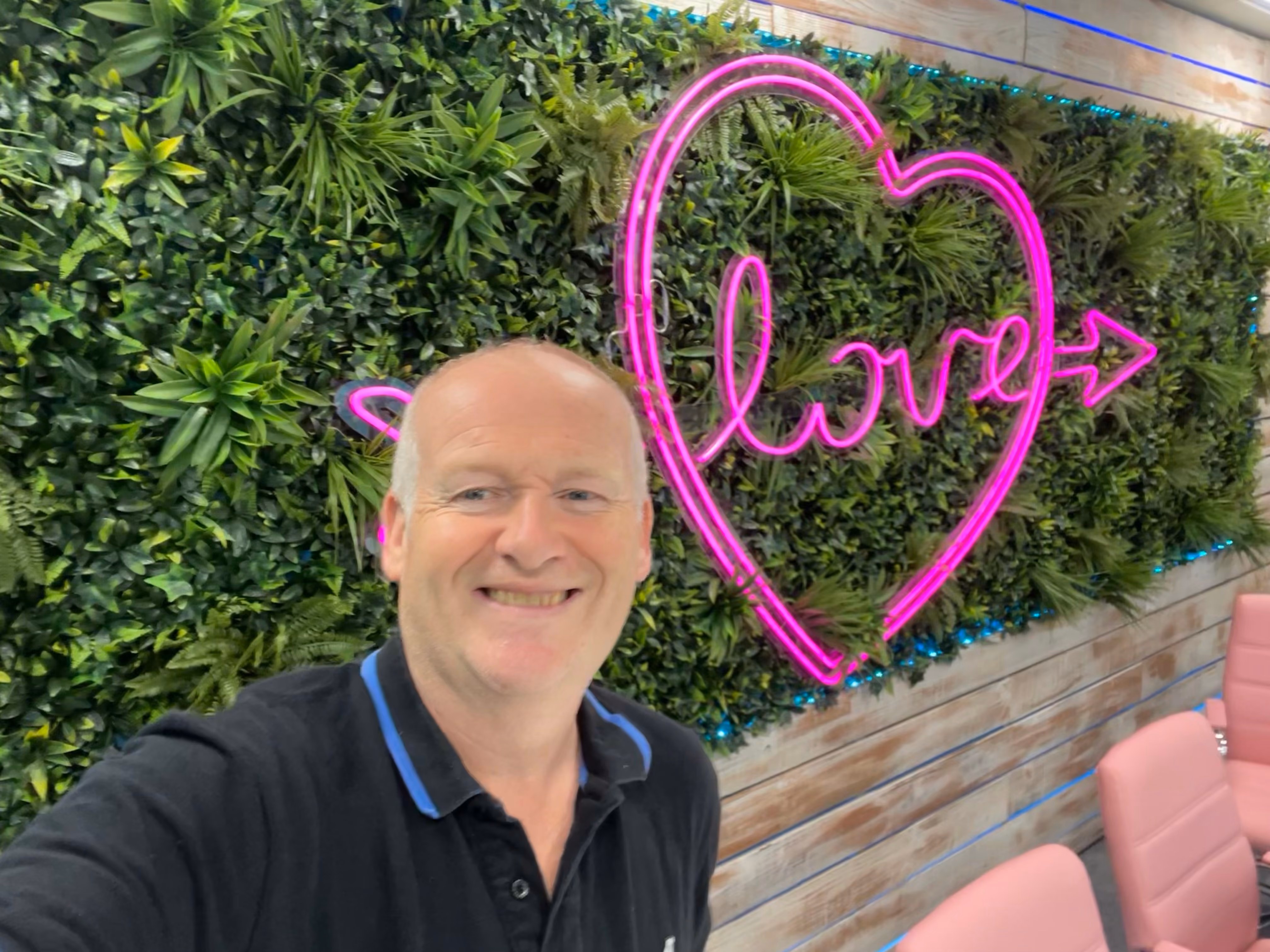 Smiling man in black shirt taking selfie in front of green plant wall with pink neon sign saying love.