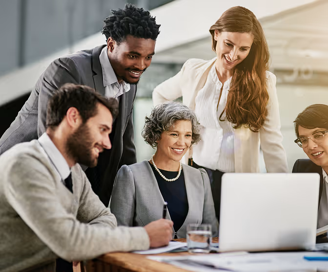 A diverse group of five business professionals gathered around a laptop, collaborating and smiling in a modern office.