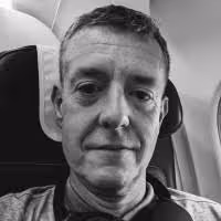 Smiling young man with short brown hair and beard wearing a dark jacket and light shirt.