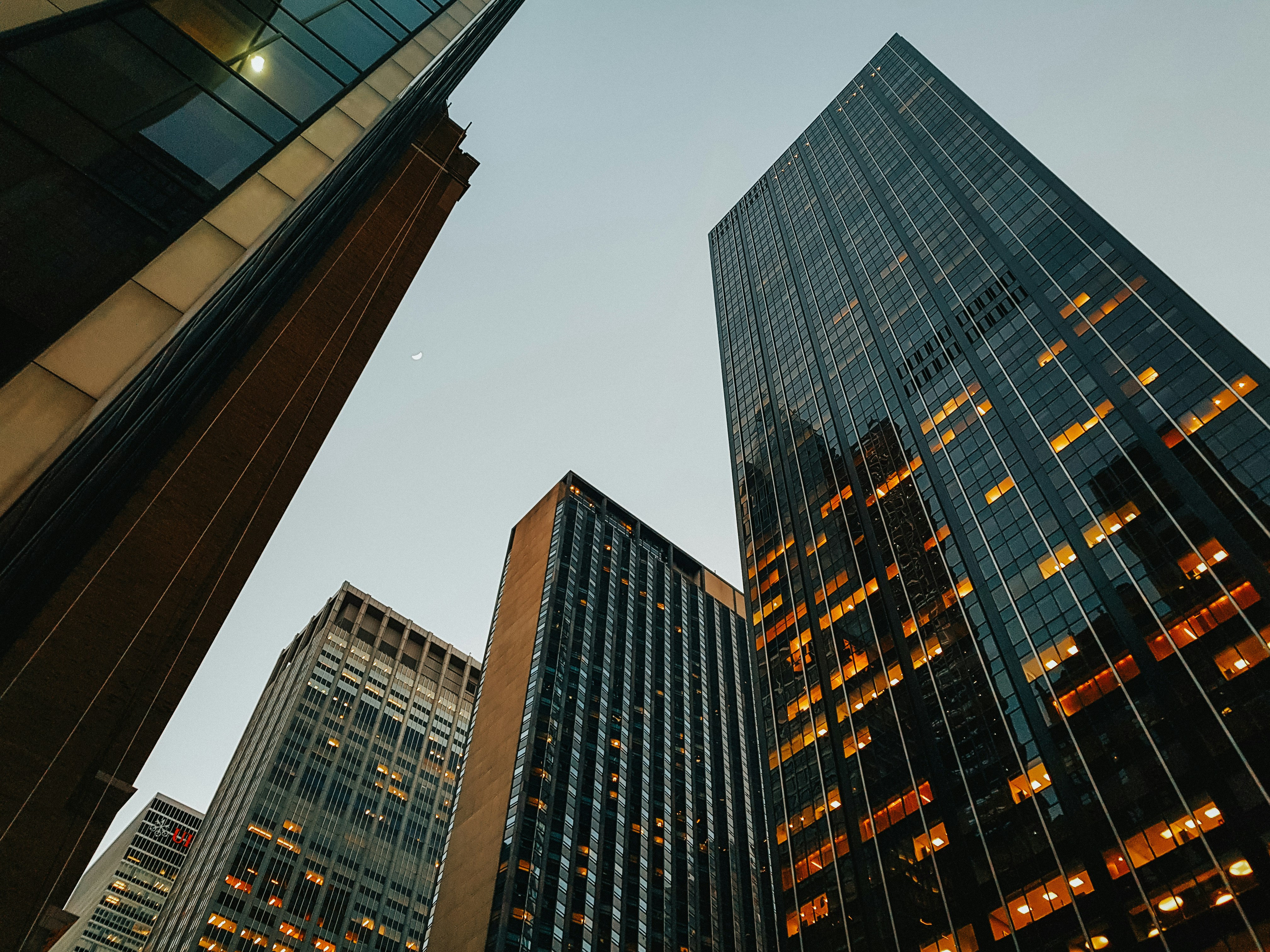 View looking up at tall skyscrapers with illuminated windows against a clear evening sky.