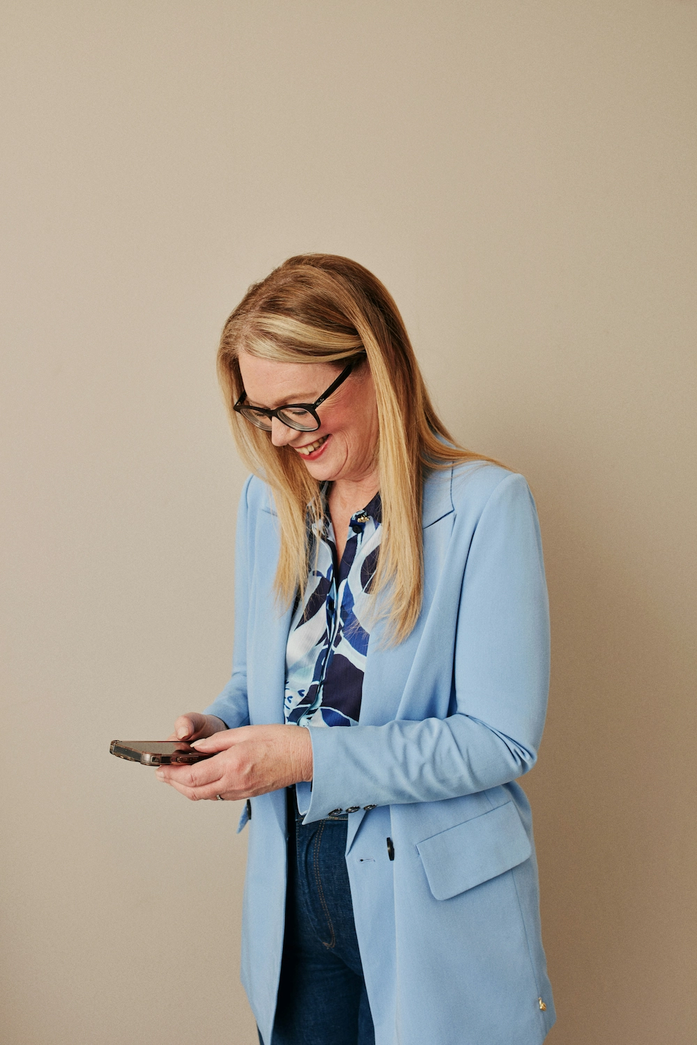 Smiling woman in a light blue blazer and patterned blouse looking at her smartphone.