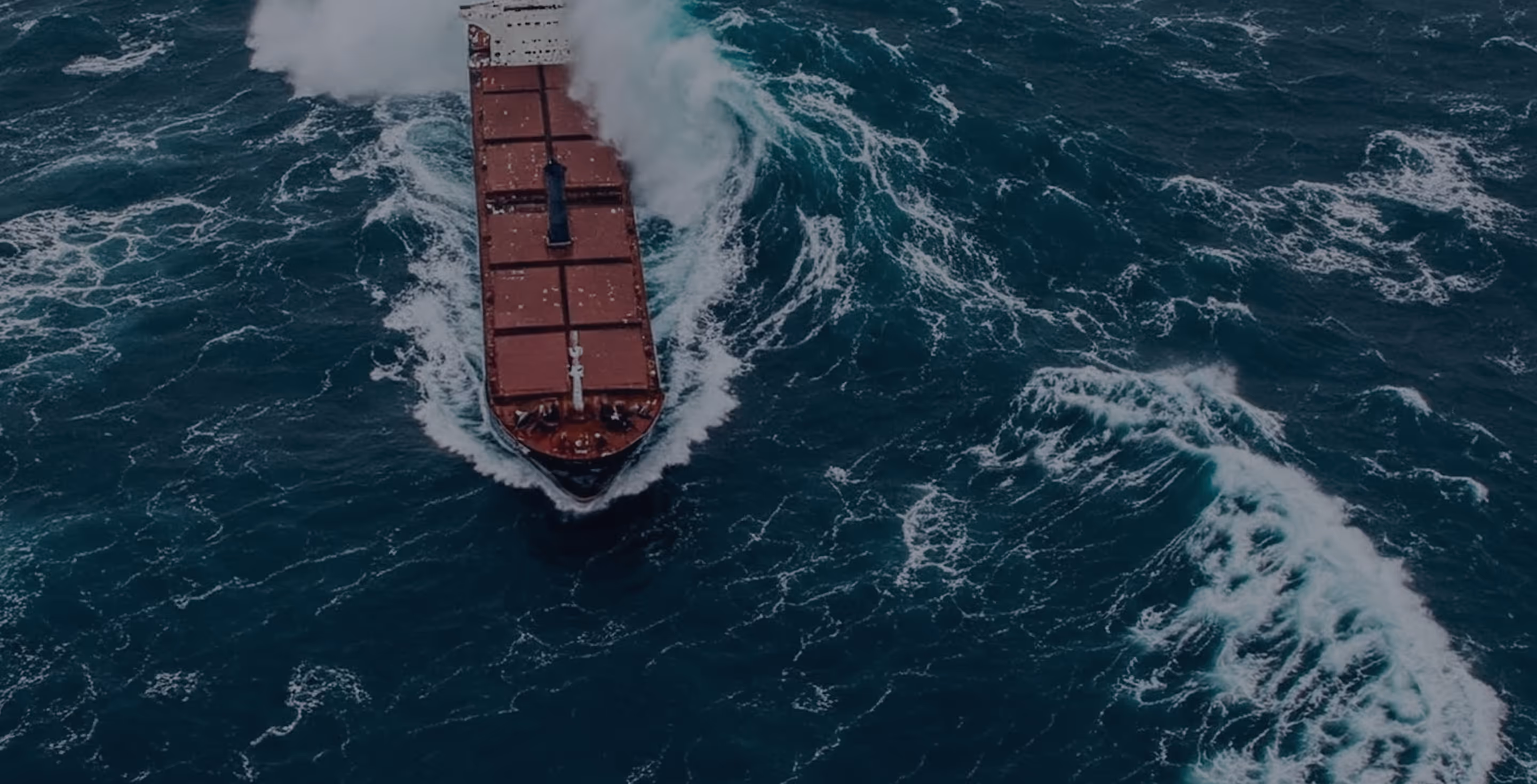 A large cargo ship navigating through rough ocean waters with waves breaking against its bow.