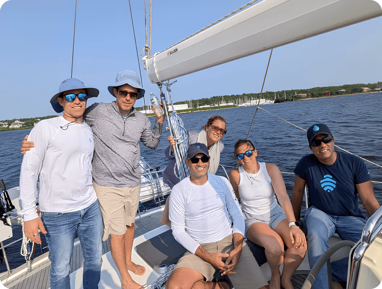 Group of six people wearing casual summer clothes and sunglasses, smiling on a sailboat with water and land with marina in the background.