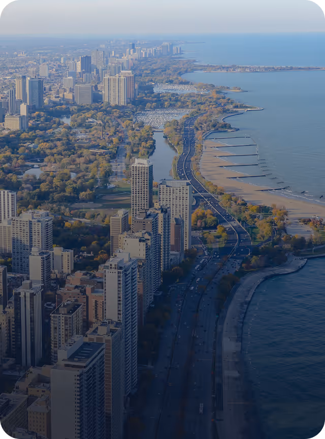 Aerial view of a coastal city with tall buildings, a highway along the beach, and boats docked in a marina.