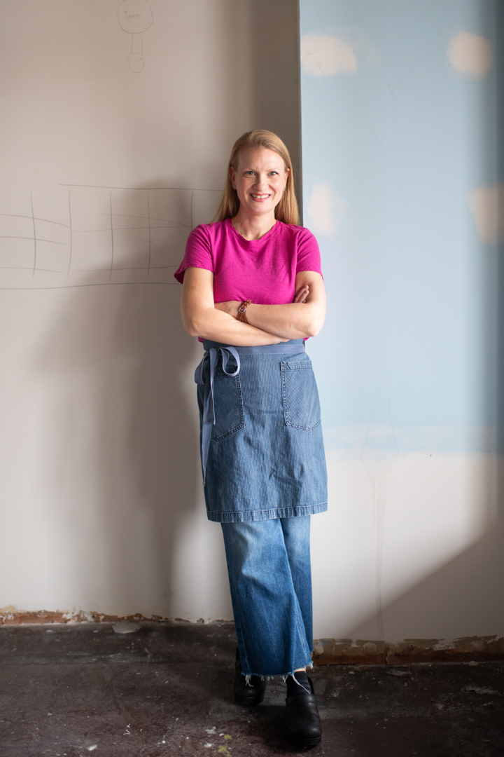 Smiling woman standing with arms crossed wearing a pink shirt and blue denim apron in a room with unfinished walls.