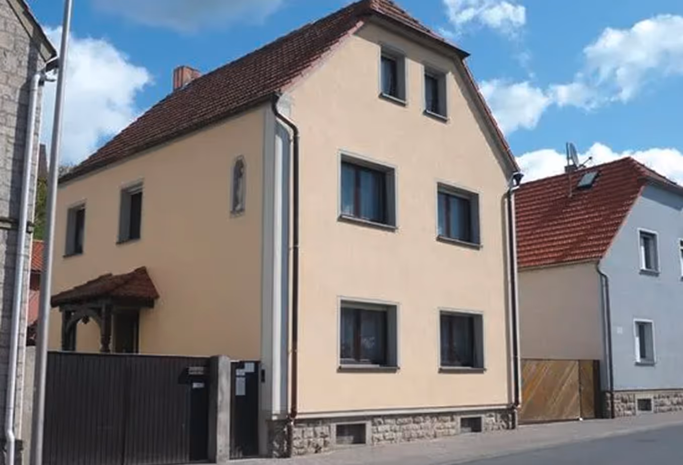 Zweistöckiges beige Wohnhaus mit rotem Ziegeldach und mehreren Fenstern an einer ruhigen Straße unter blauem Himmel mit Wolken.