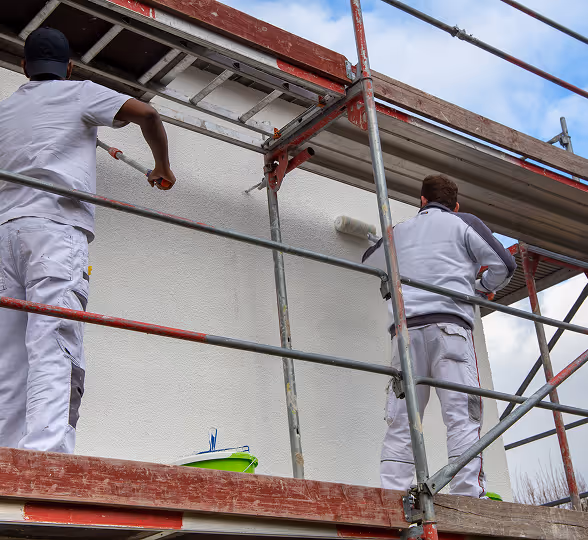 Zwei Maler in weißer Arbeitskleidung streichen eine weiße Außenwand von einem Gerüst aus.