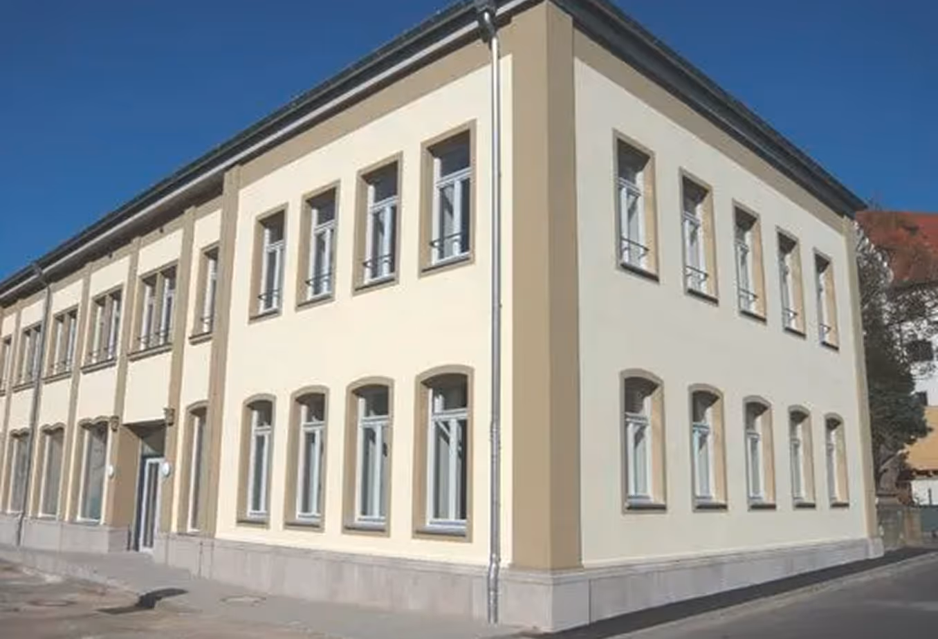 Eckansicht eines zweistöckigen, hellen Gebäudes mit mehreren symmetrischen Fenstern unter blauem Himmel.